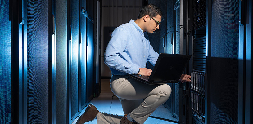 A man kneeling on the ground next to a column of HPC servers, with a laptop resting on his right knee.